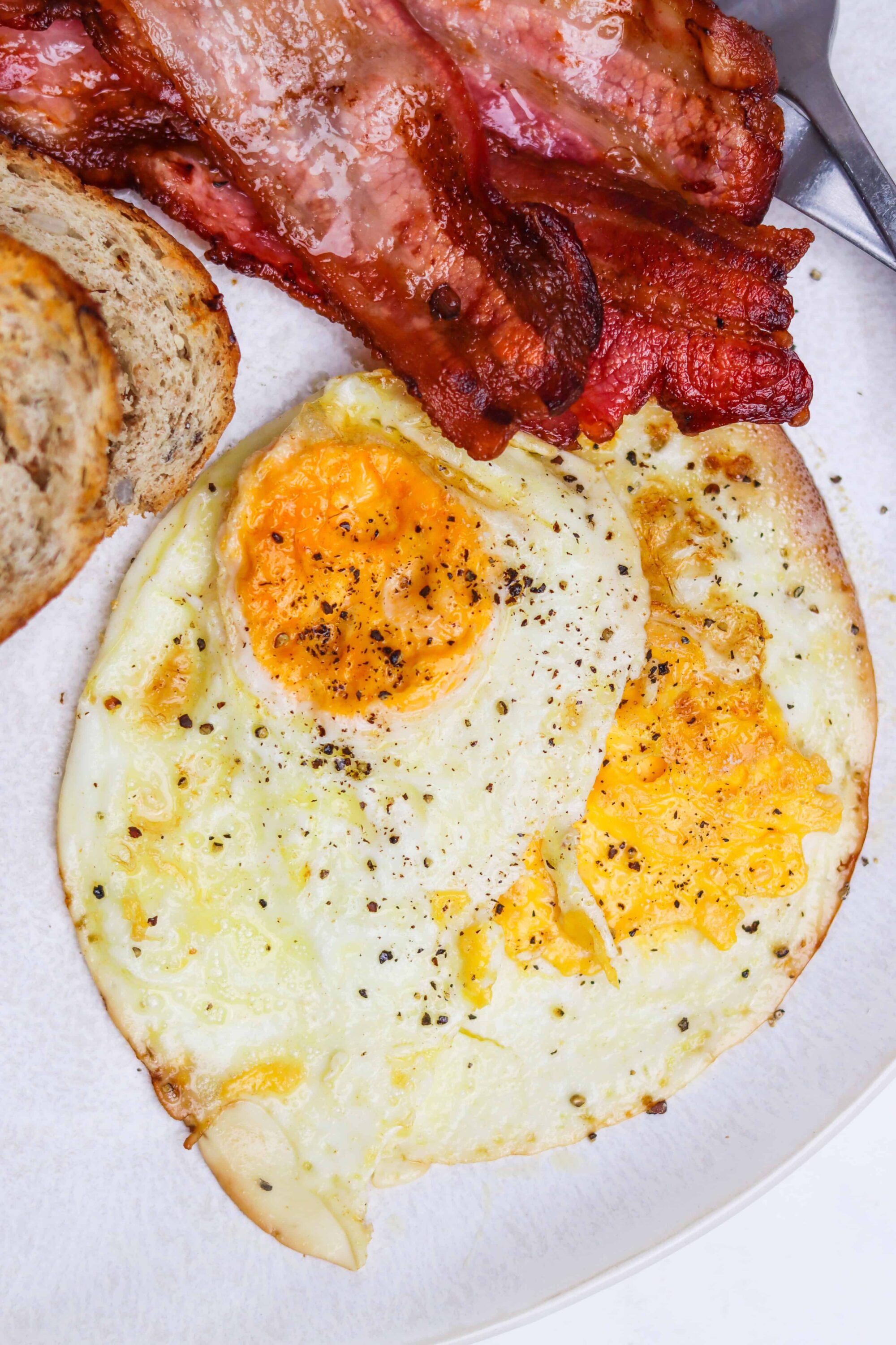 Crispy bacon, sunny side up eggs, whole grain toast on white plate.