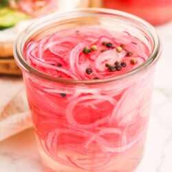 Pickled red onions in a glass jar with peppercorns, bright pink color.