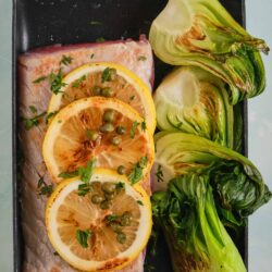 Juicy roasted lemon fish with charred lettuce and fresh herbs in a black roasting pan.