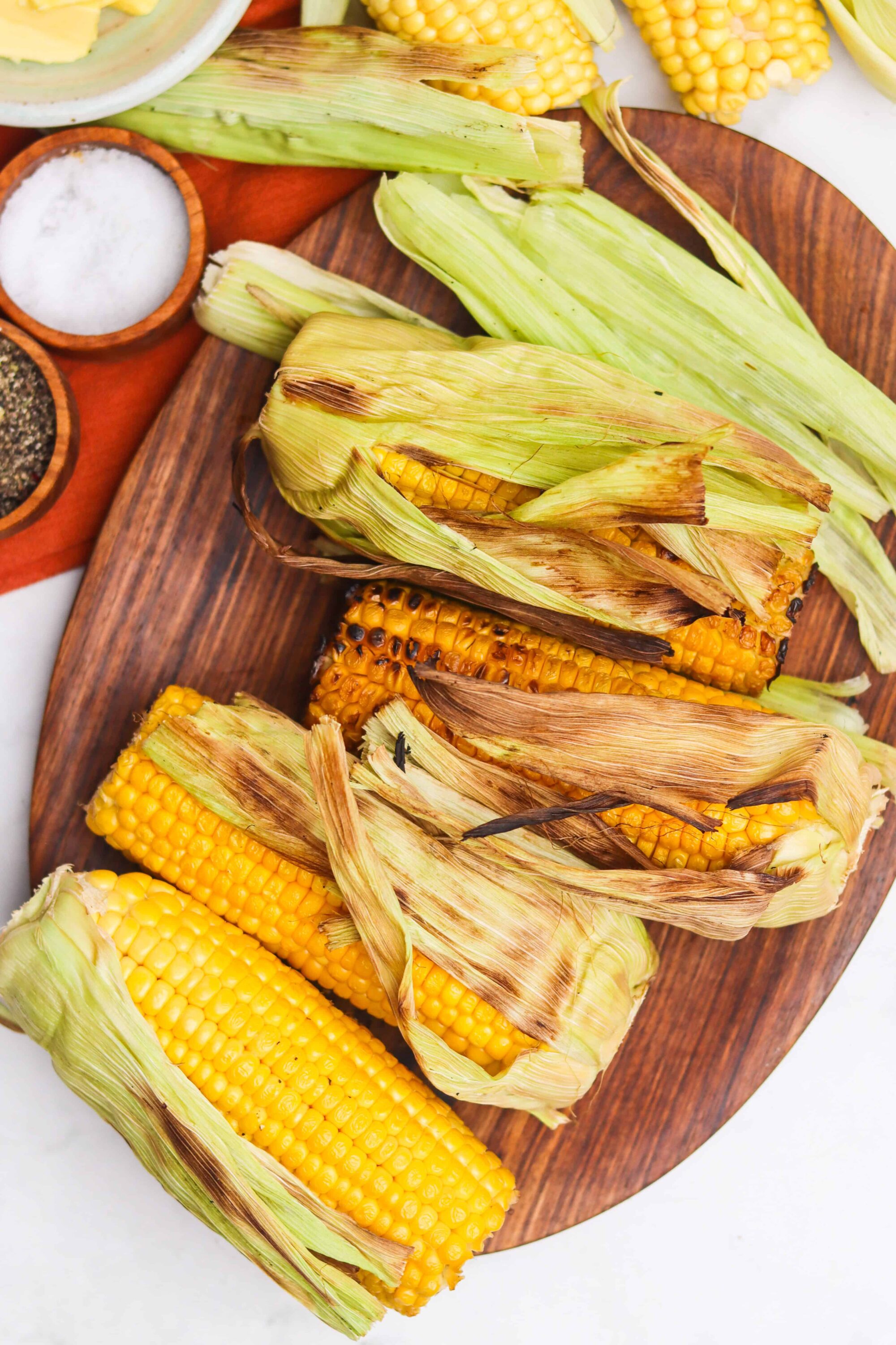 Grilled corn on the cob with charred husks on a wooden serving platter.