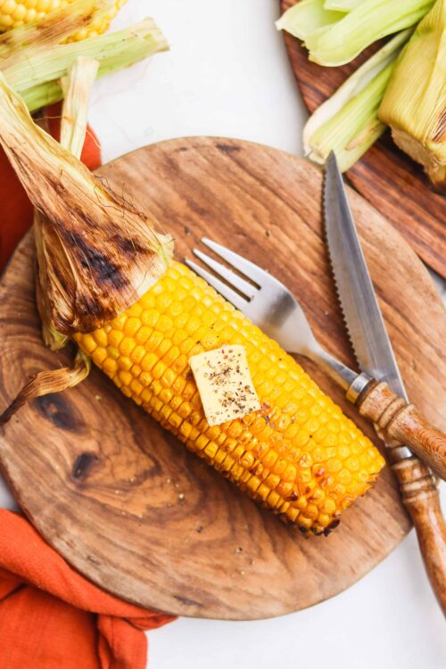 Corn on the cob with butter, grilled corn, summer vegetables, wooden cutting board, kitchen knife and fork.