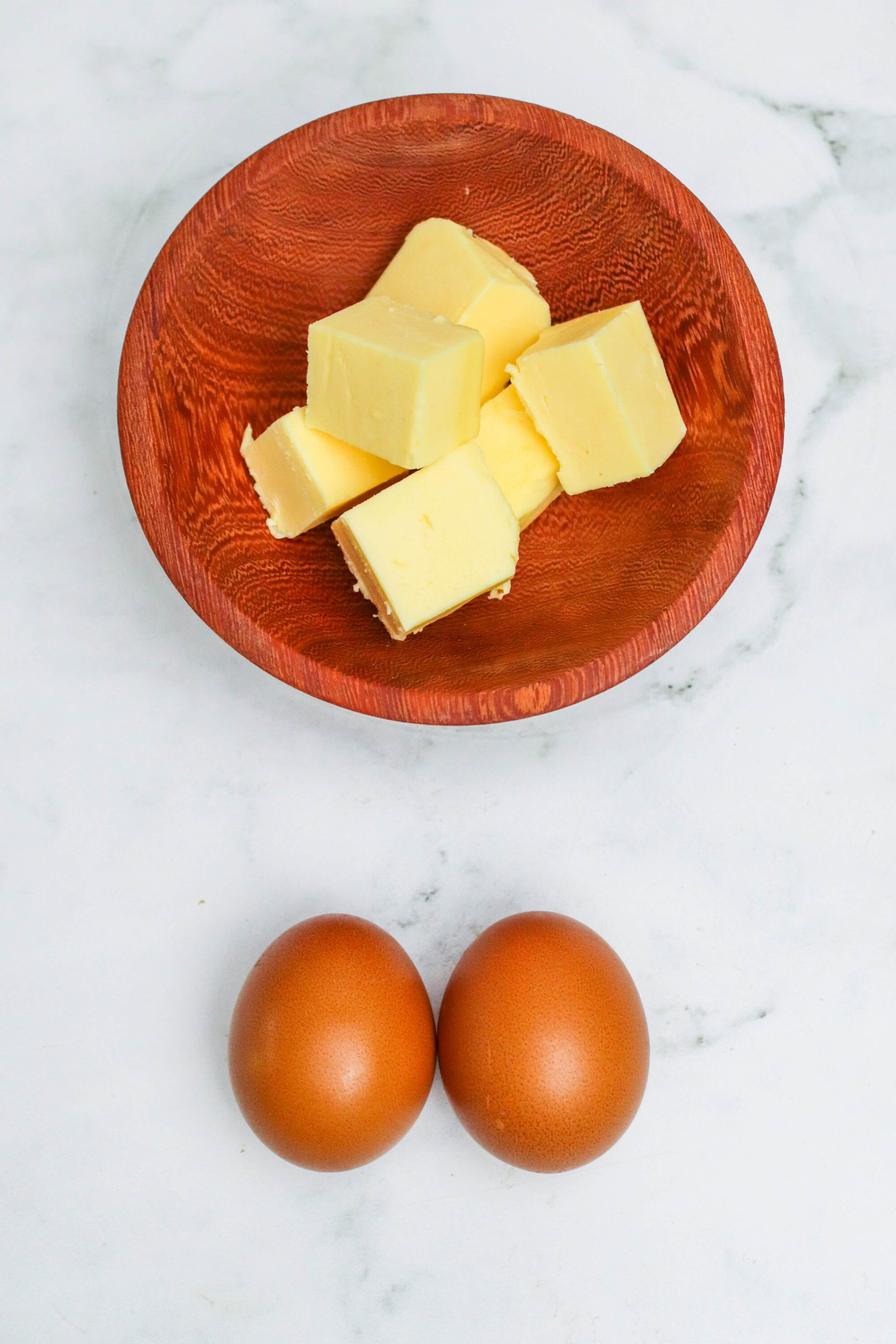 Buttery white chocolate chunks in a wooden bowl with two brown eggs on a white marble surface.