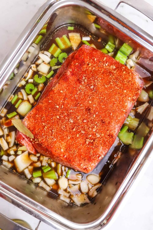 Juicy pot roast with seasoning, cooking in a stainless steel roasting pan with vegetables.