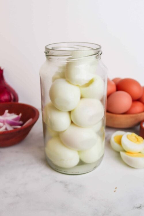 Boiled eggs in a glass jar with fresh eggs and vegetables in the background.