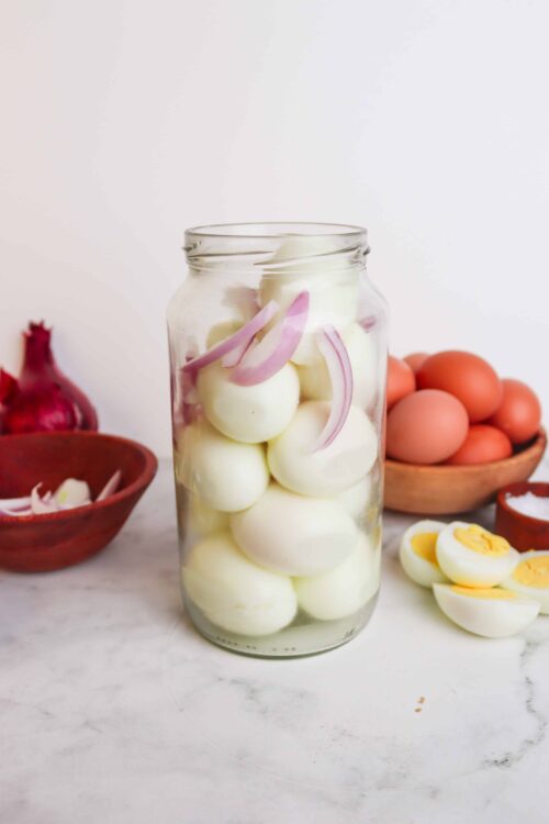 Pickled hard-boiled eggs with onions in a glass jar on white background.