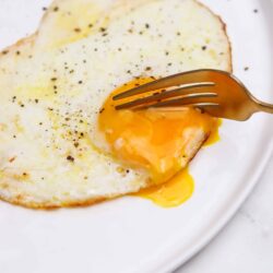Fluffy over-easy egg with runny yolk on white plate, black pepper seasoning, close-up, breakfast recipe.