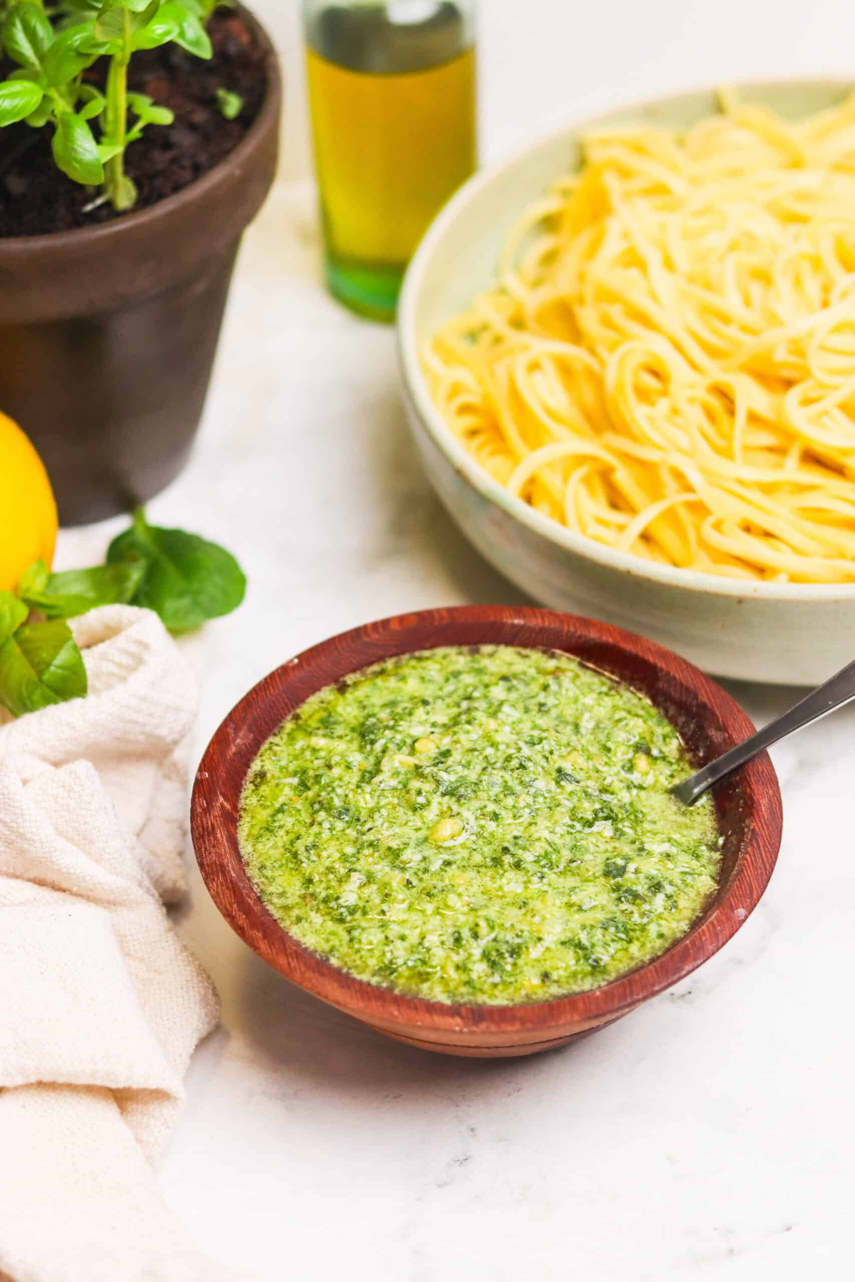 Creamy pesto sauce in a wooden bowl with spaghetti, fresh basil, olive oil, and lemon on a white marble surface.