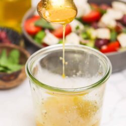 Rich honey being drizzled from a spoon into a glass jar, with fresh salad ingredients in the background.