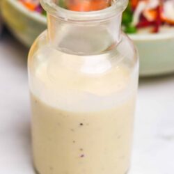 Creamy salad dressing in a glass bottle with a colorful vegetable salad in the background.