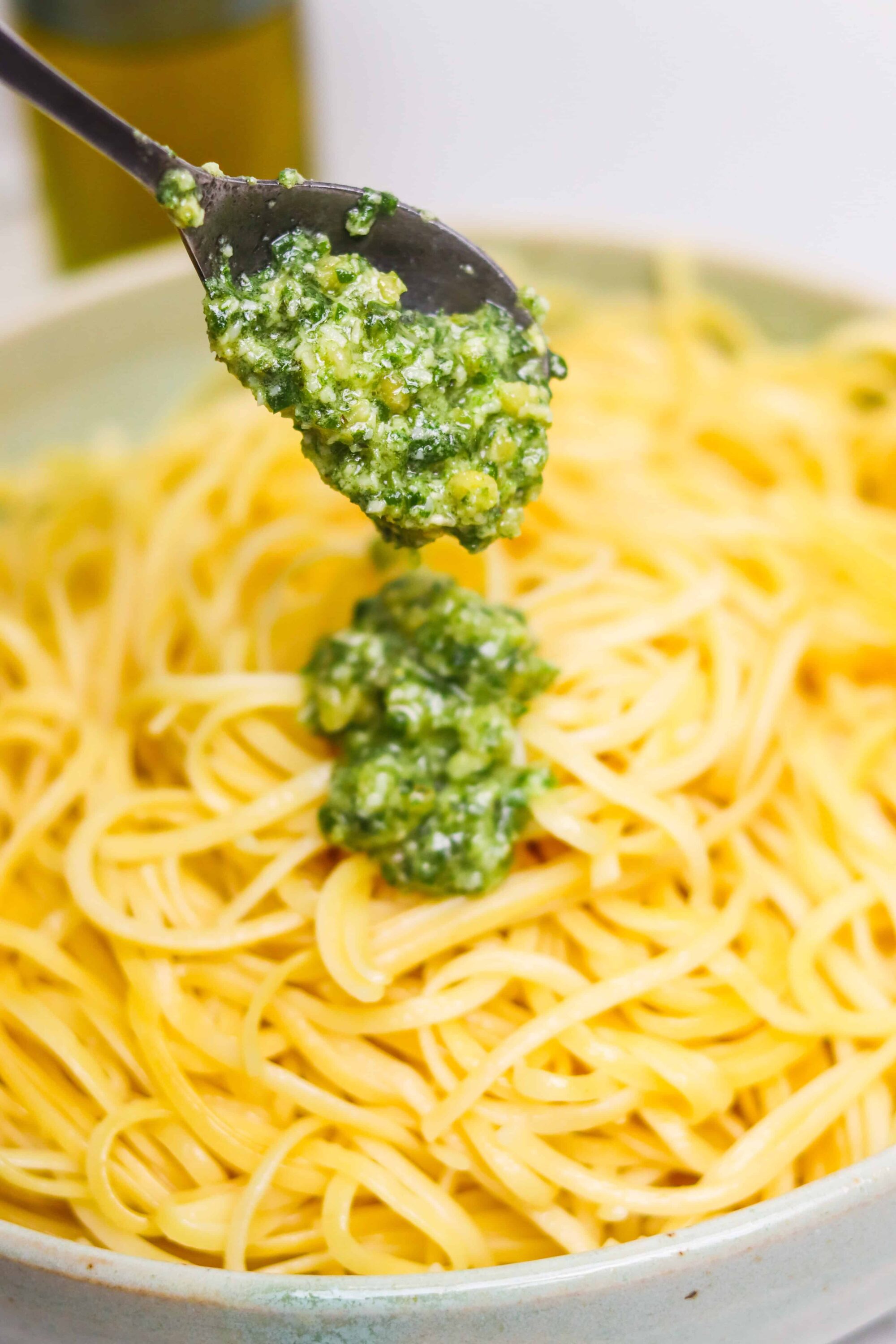 Creamy basil pesto being added to cooked spaghetti in a white bowl.
