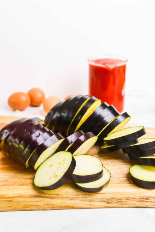 Sliced eggplant on wooden cutting board with eggs and tomato sauce in background.