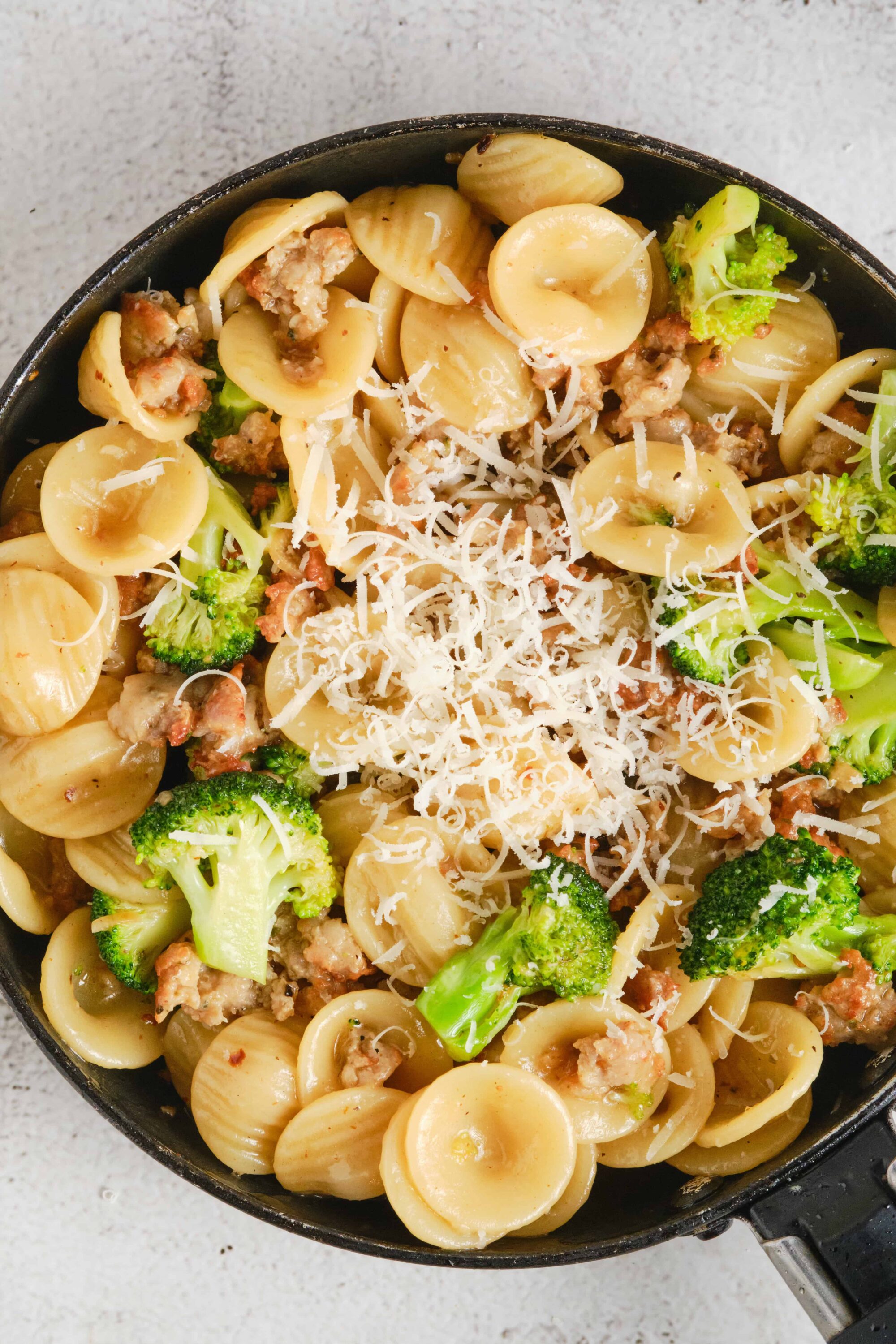 Creamy pasta with broccoli and ground meat in a skillet, topped with shredded cheese.