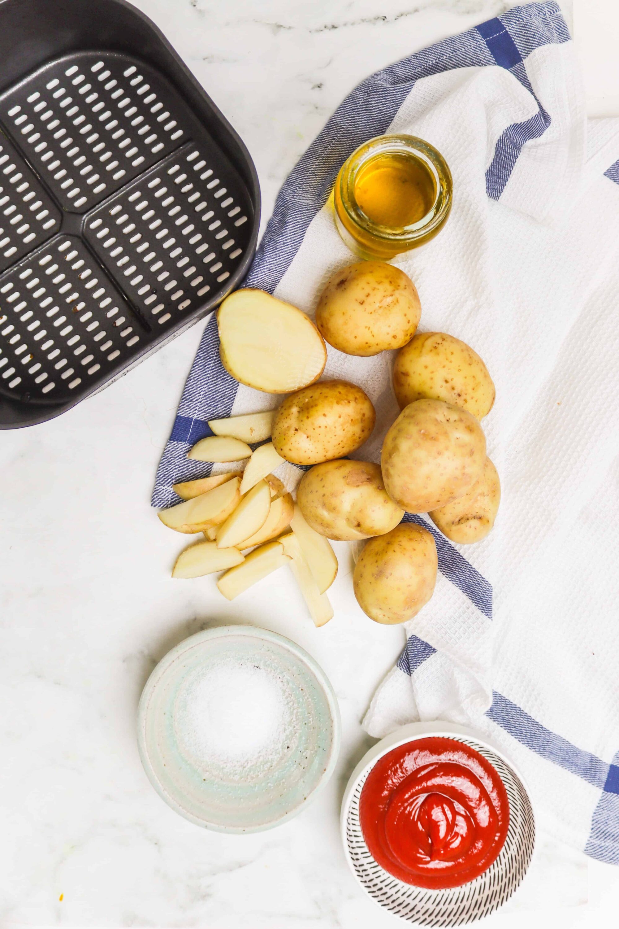 Fresh potatoes, garlic, olive oil, salt, and ketchup on a white marble surface for baking or cooking.