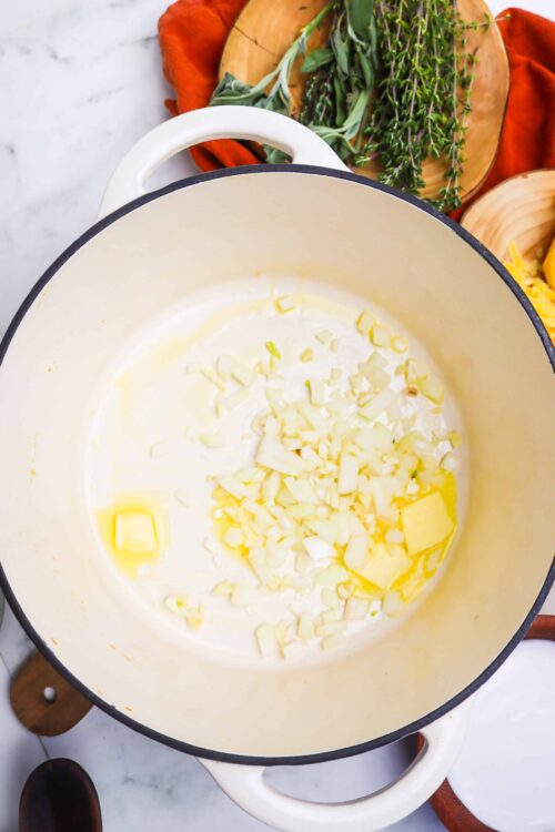 Butter and chopped onions melting in a white cast iron skillet with fresh herbs and wooden cutting board in background.