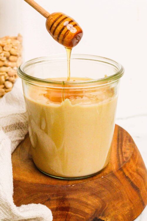 Pure honey being drizzled into a jar of creamy honey spread on a wooden surface.