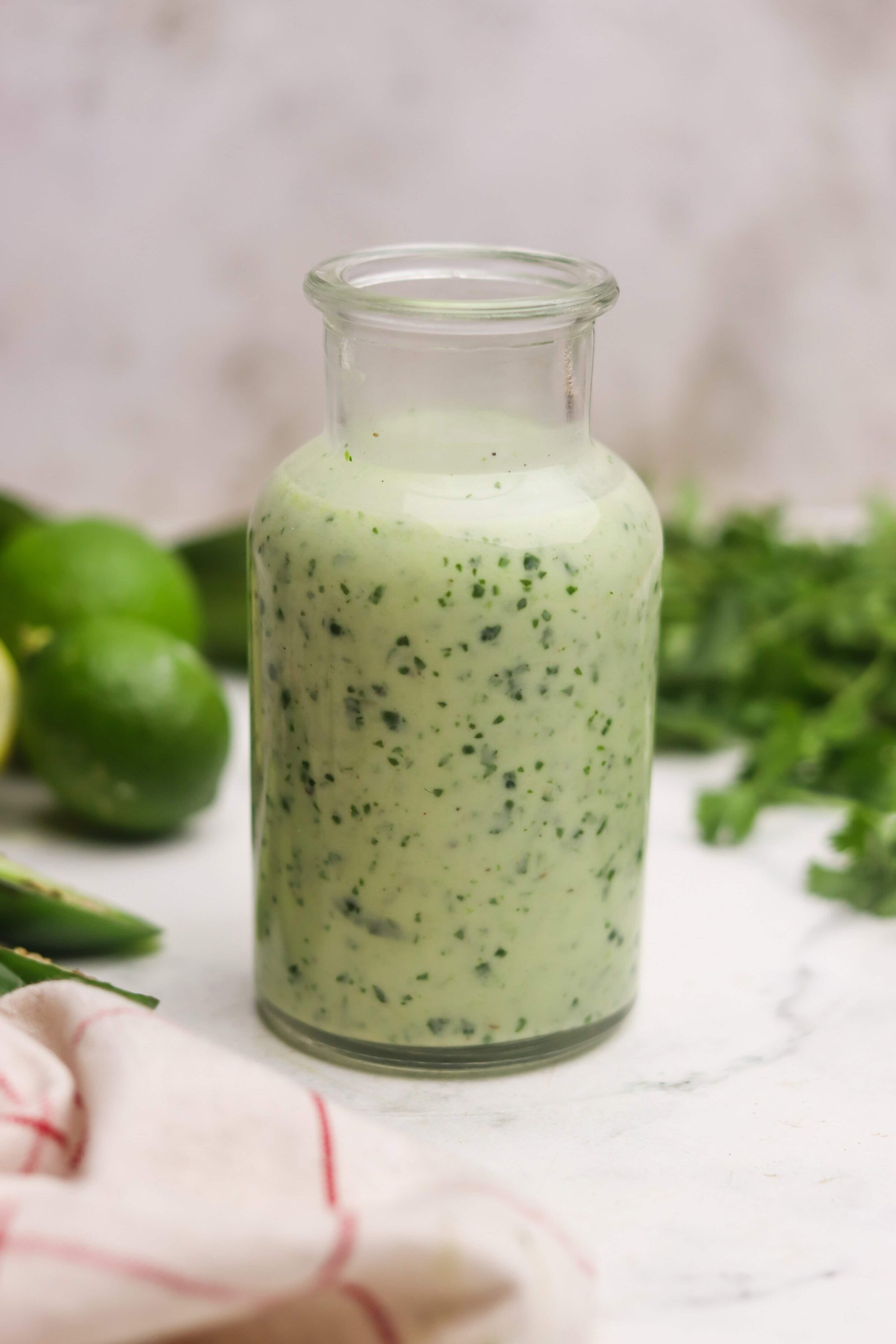 Creamy cilantro lime dressing in a glass jar on a white surface with fresh limes and herbs.