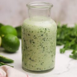 Creamy cilantro lime dressing in a glass jar on a white surface with fresh limes and herbs.