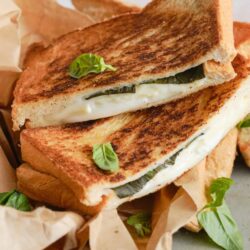 Golden brown grilled cheese sandwich with melted cheese and basil on parchment paper.