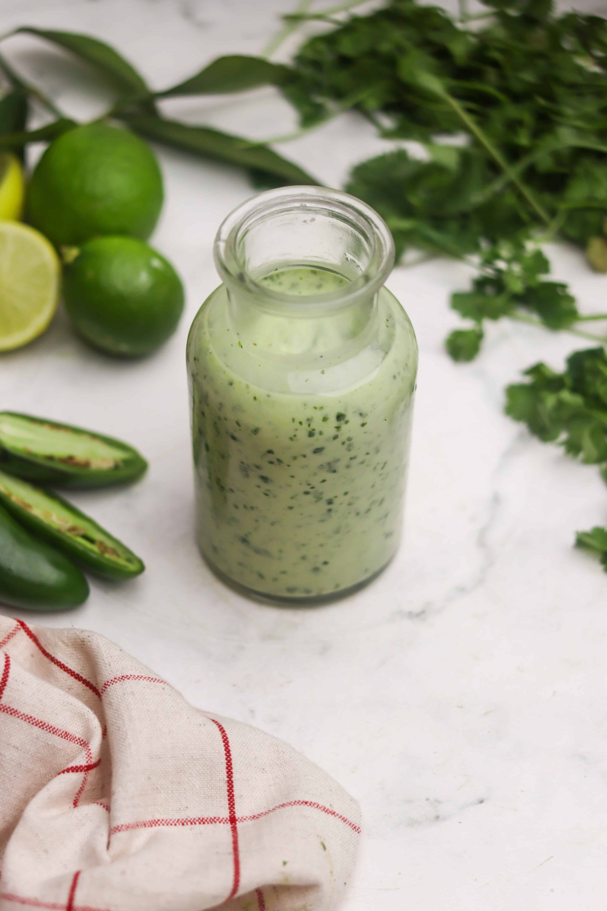 Lime cilantro dressing in a glass bottle with fresh limes and herbs surrounding it.