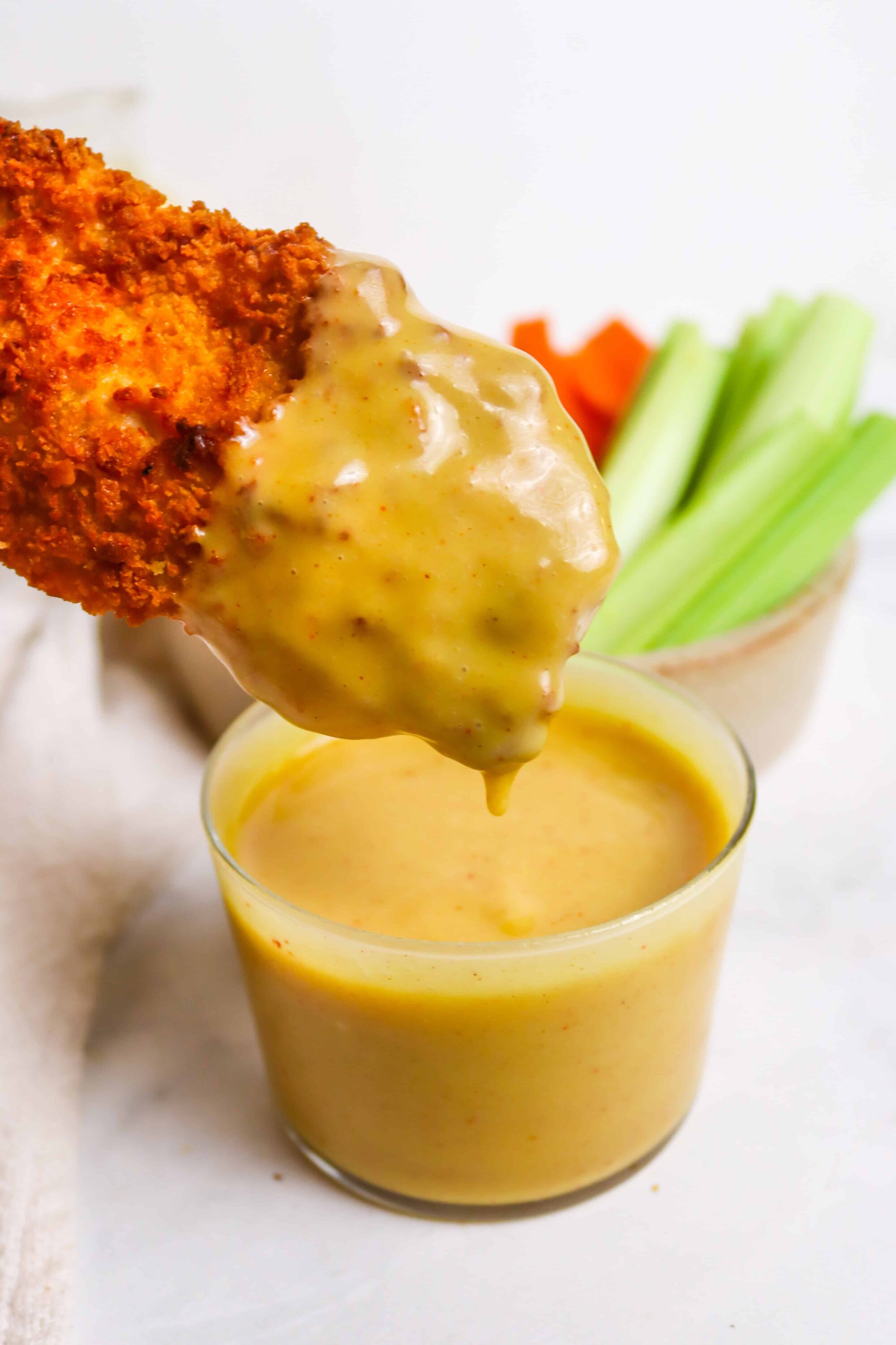 Crispy fried chicken tender with creamy mustard sauce dipping, fresh veggie sticks in the background.