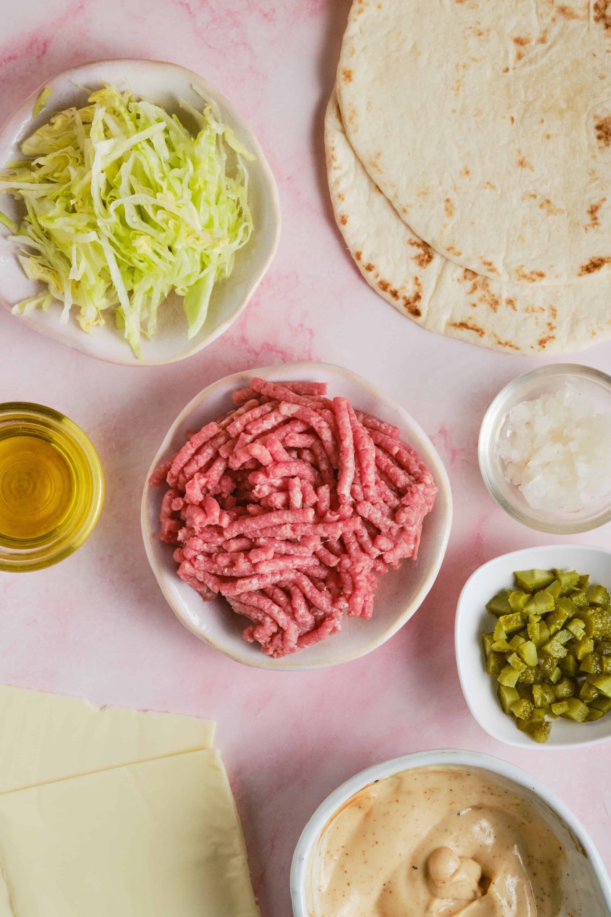 Ground beef, shredded lettuce, flour tortillas, chopped pickles, cheese slices, onions, and sauce for taco preparation.