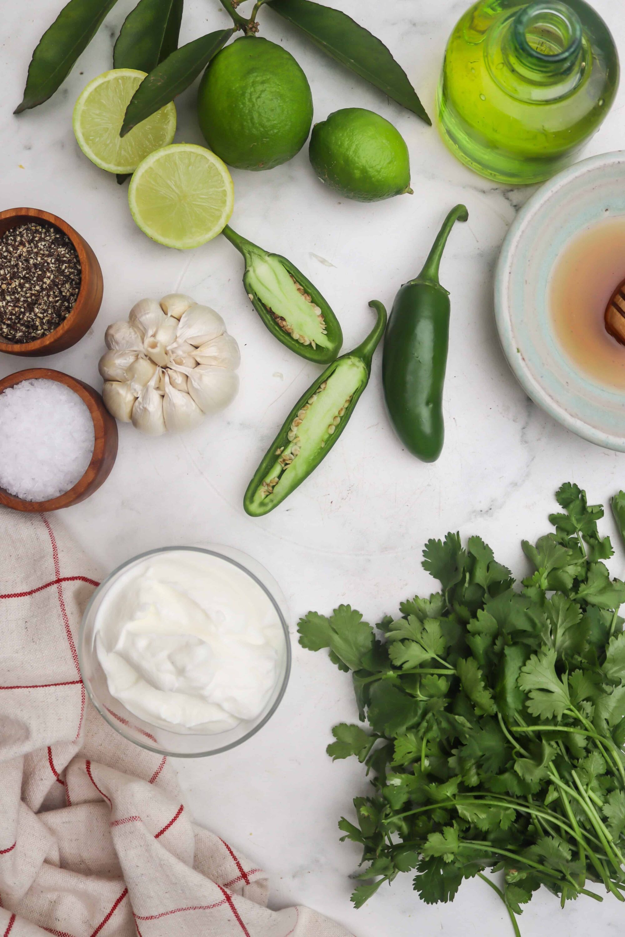 Fresh limes, garlic, jalapeño peppers, salt, pepper, olive oil, cilantro, and yogurt on a white marble surface for spicy, flavorful cooking.