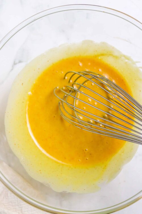 Creamy yellow custard mixture for baking or pastry filling in a clear glass bowl with whisk.