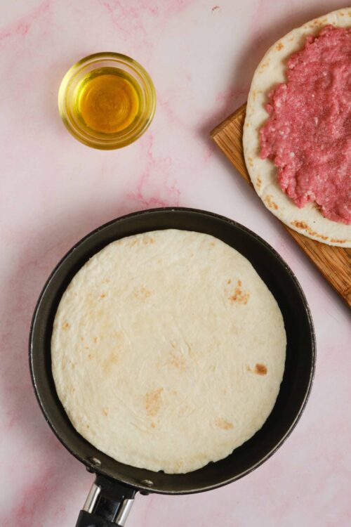 Fluffy pizza dough in a skillet with toppings and olive oil on a pink marble surface.