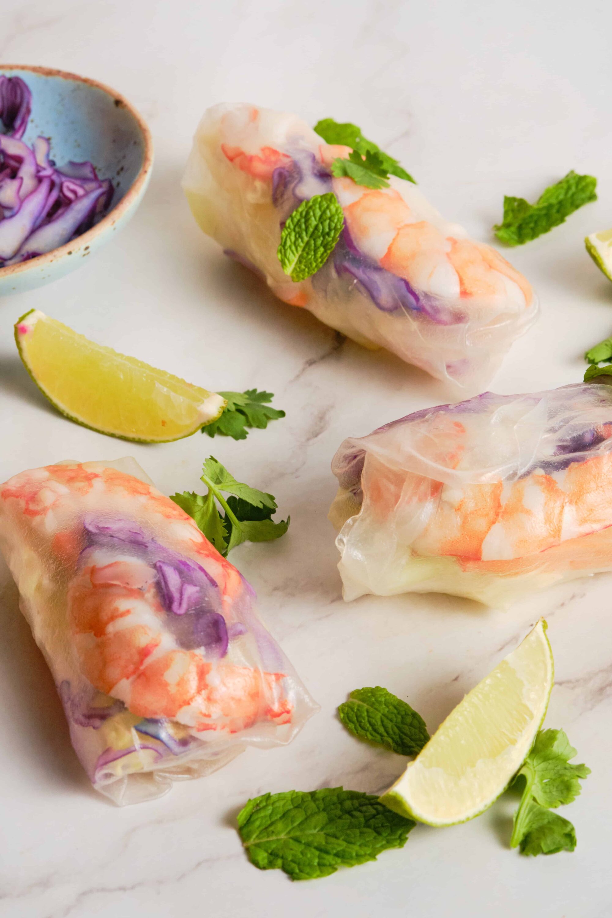 Fresh spring rolls with shrimp and vegetables on a white marble surface, garnished with lime and mint.