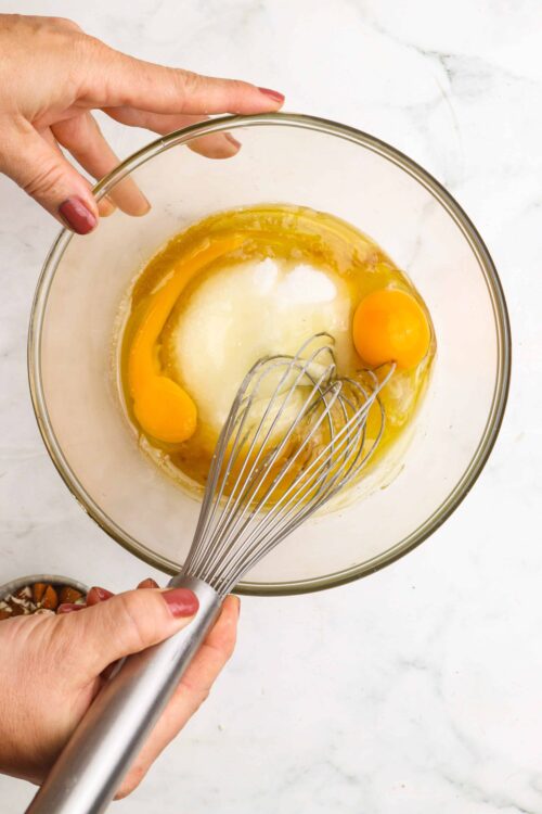 Creamy egg mixture for baking recipes, whisked in a glass bowl with hands adding ingredients.