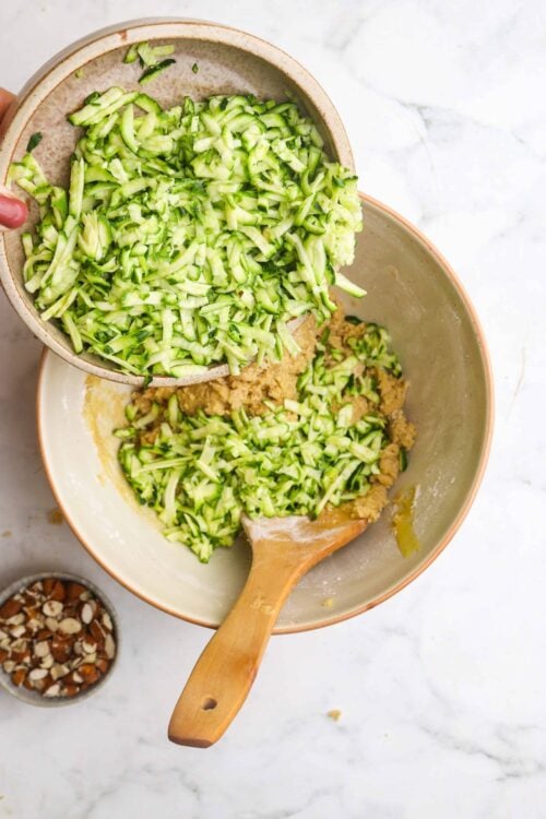 Chopped zucchini being added to a mixing bowl with cookie dough, preparing for zucchini bread or baking.