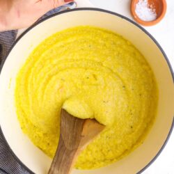 Creamy polenta cooking in a white cast-iron skillet with a wooden spoon.