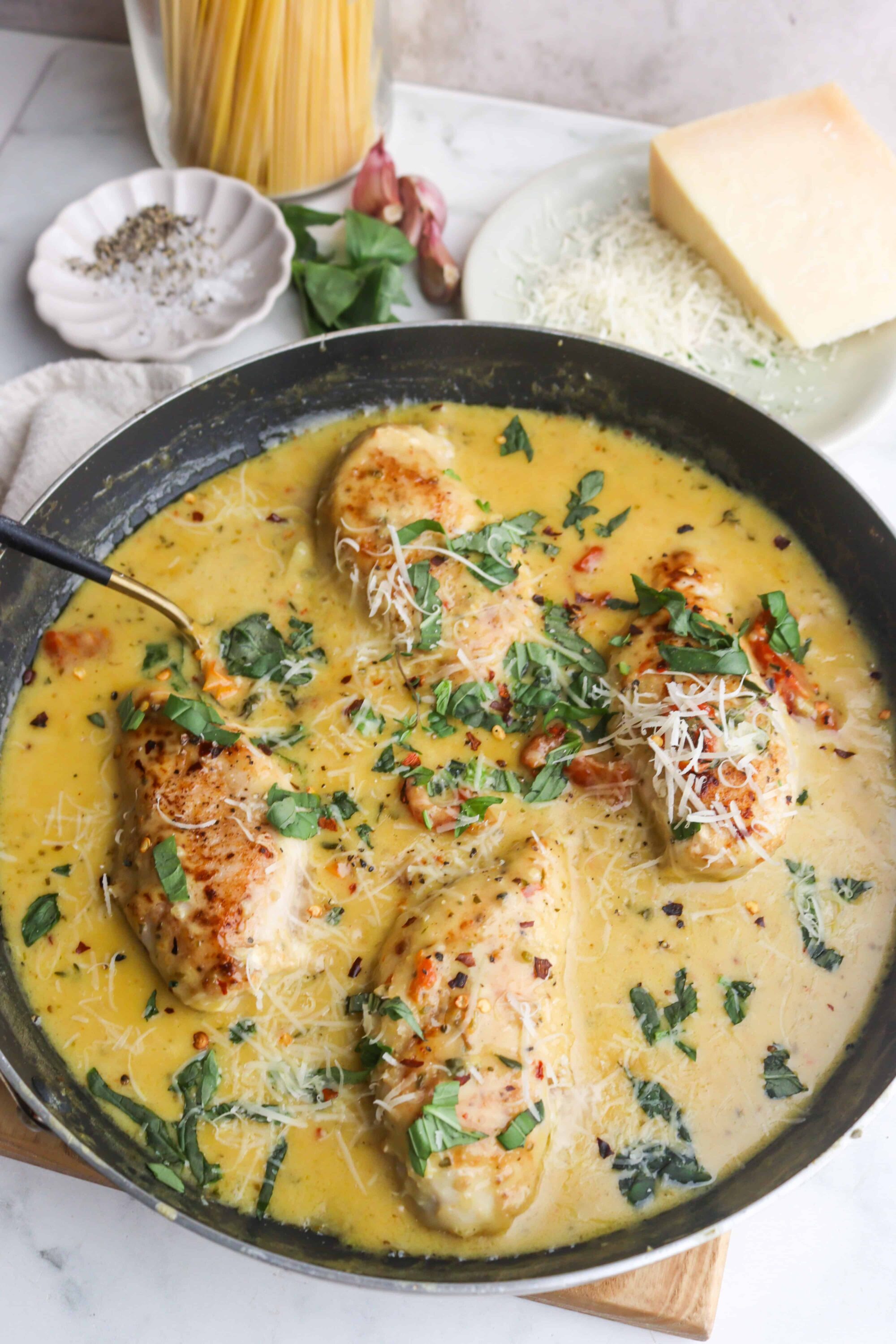 Butter chicken in a creamy sauce with herbs and cheese, cooked in a skillet, with Parmesan and garlic in the background.