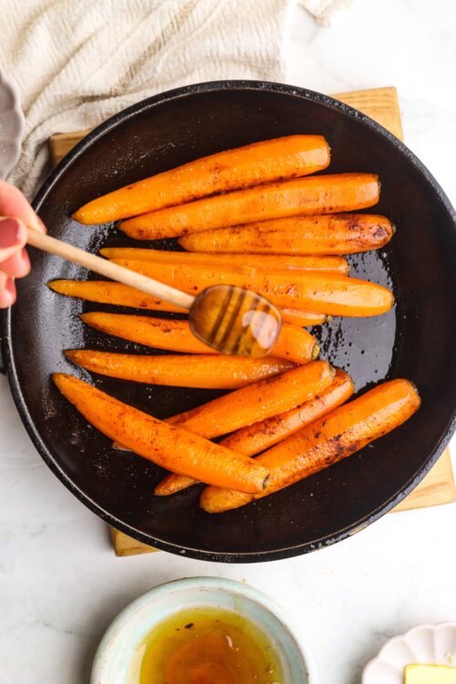 Roasted carrots cooking in a cast iron skillet with spices and honey drizzle.