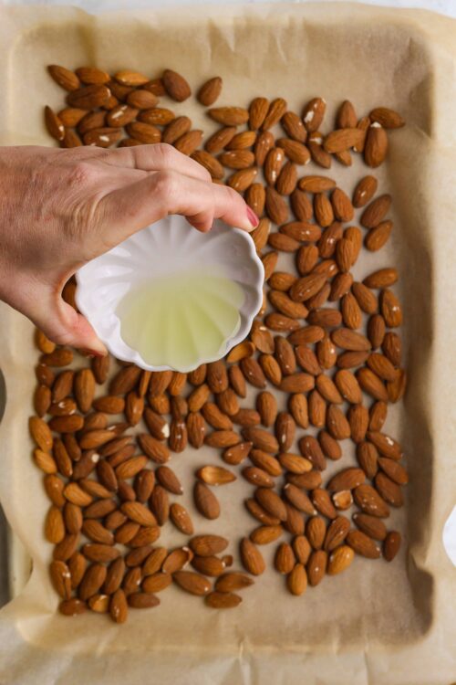 Roasted almonds on baking sheet with olive oil being added, gluten-free snack preparation.