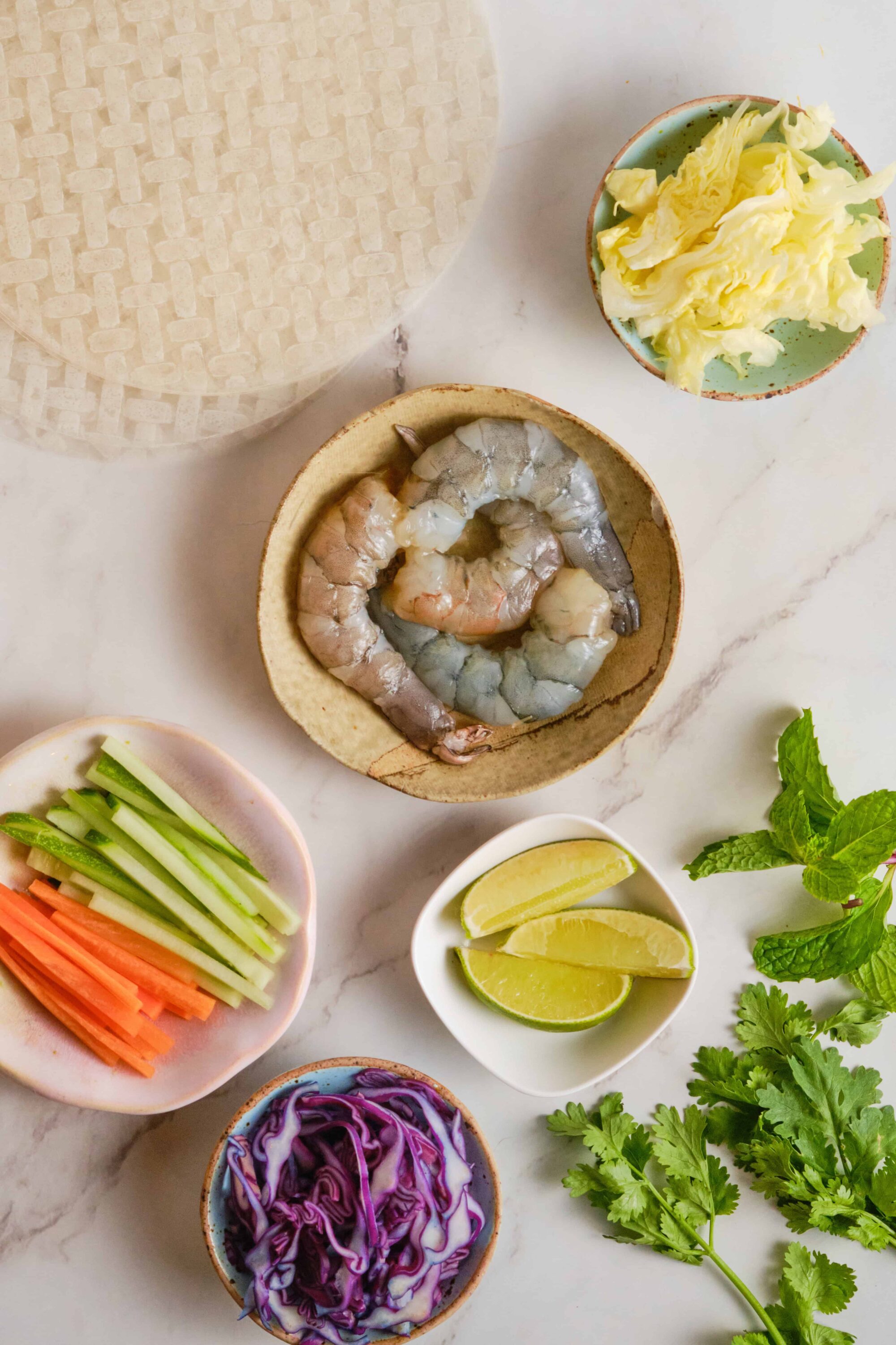 Thinly sliced vegetables, raw shrimp, lime wedges, and herbs for Vietnamese spring rolls preparation.