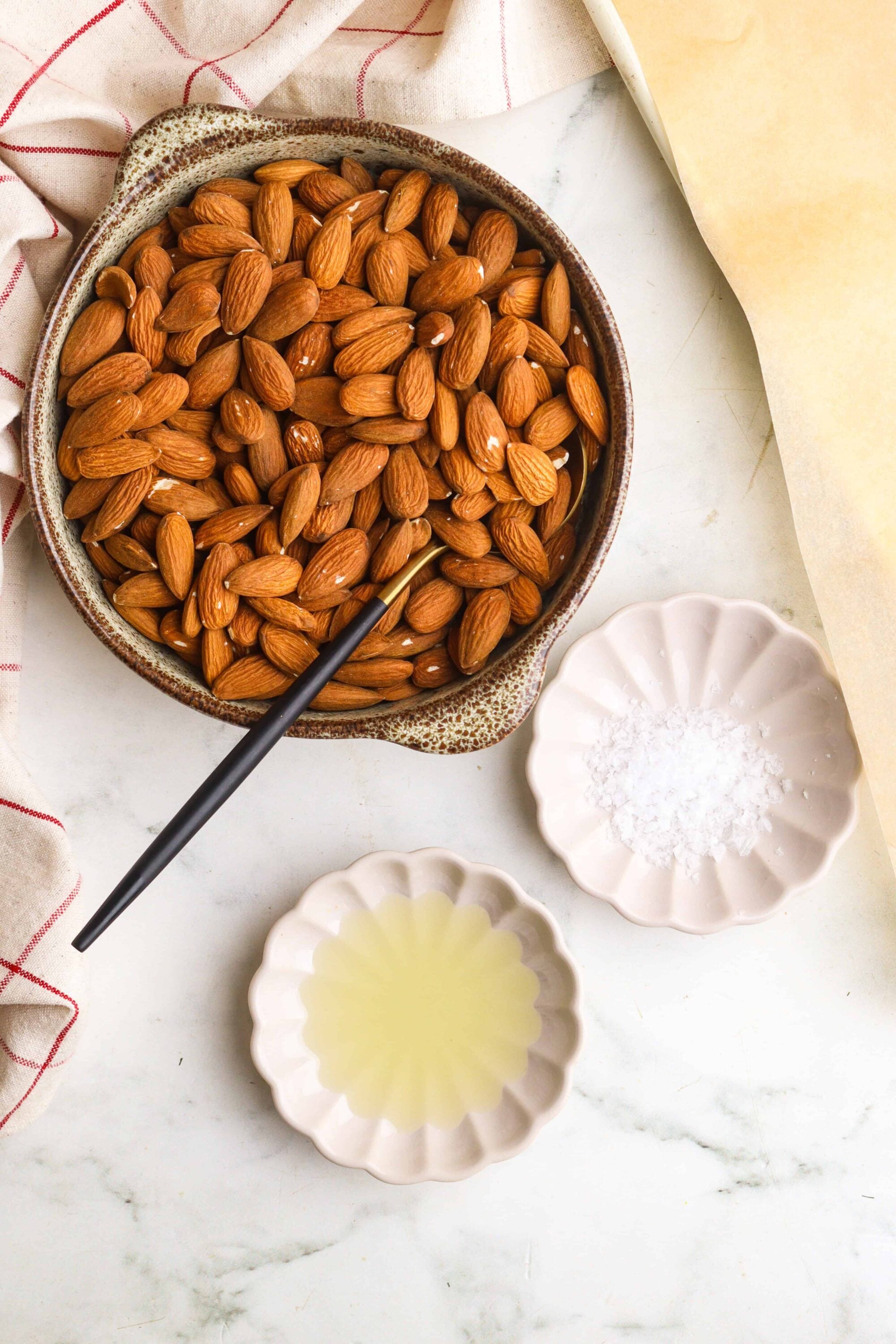 Toasted almonds in a bowl with sea salt, sweetener, and oil on a white marble surface, perfect for baking and snacking.