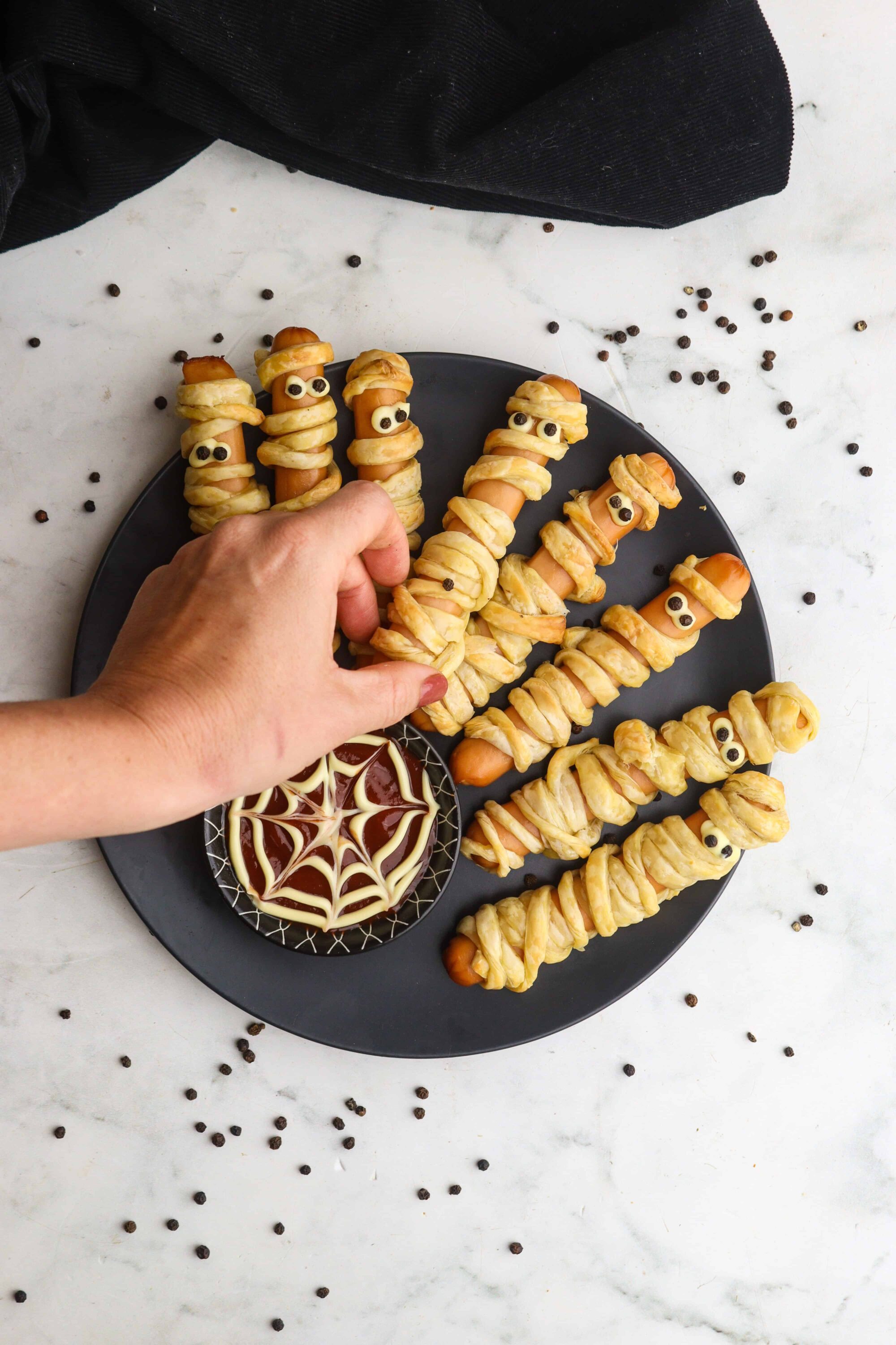 Mini hot dog mummies with twisted puff pastry and candy eyes, perfect for Halloween themed snacks. Served with spooky web dipping sauce.
