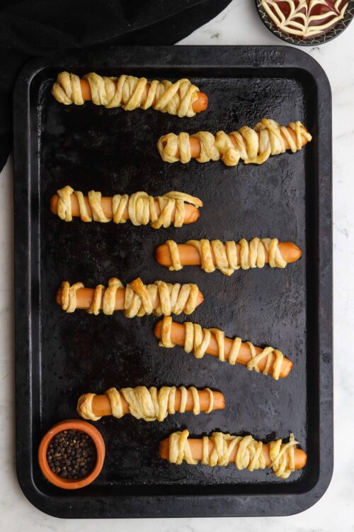 Hot dogs wrapped in puff pastry, ready for baking on a black baking sheet.