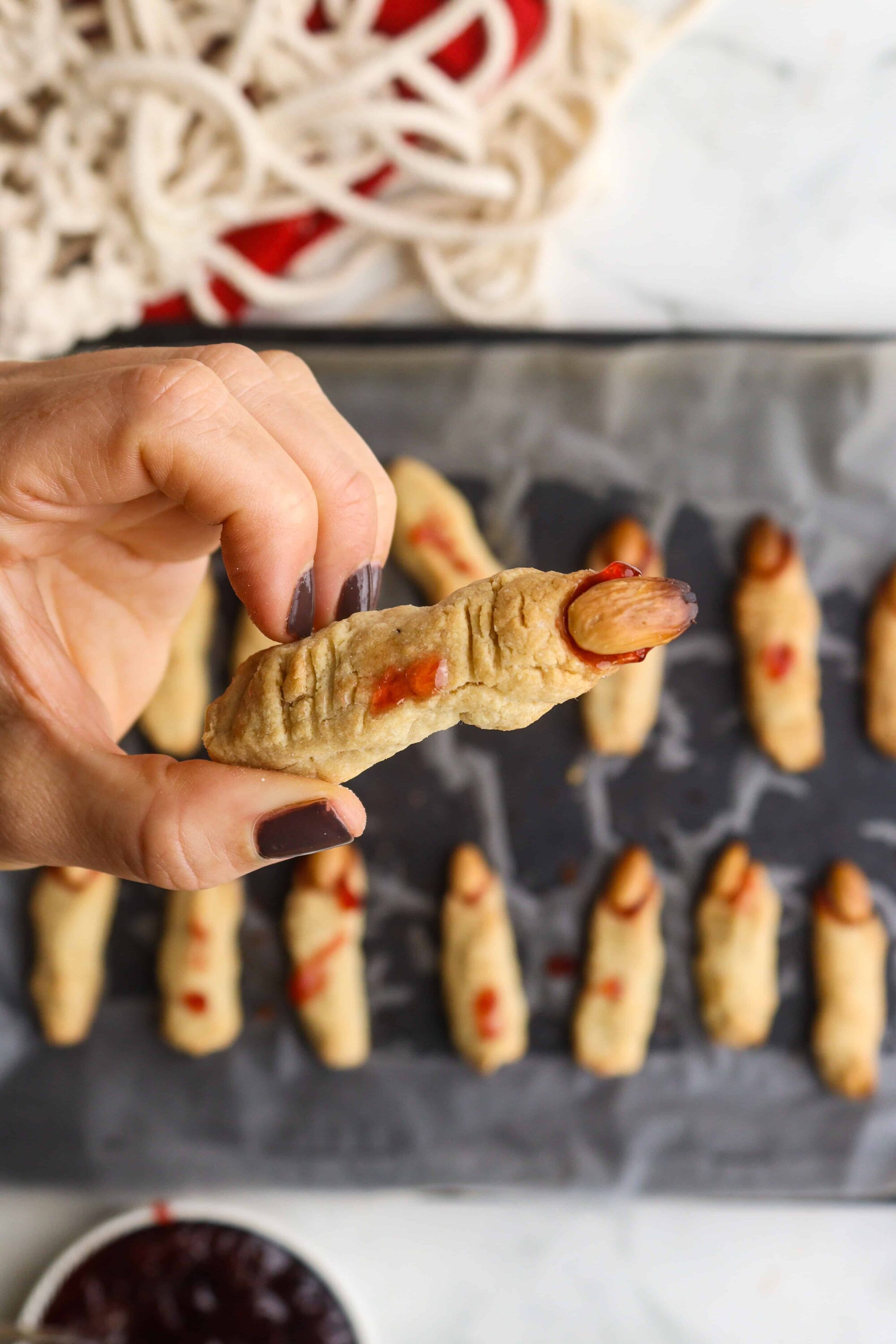 Hot dog-shaped cookie with almond, red jelly, and cookie dough, on baking sheet.