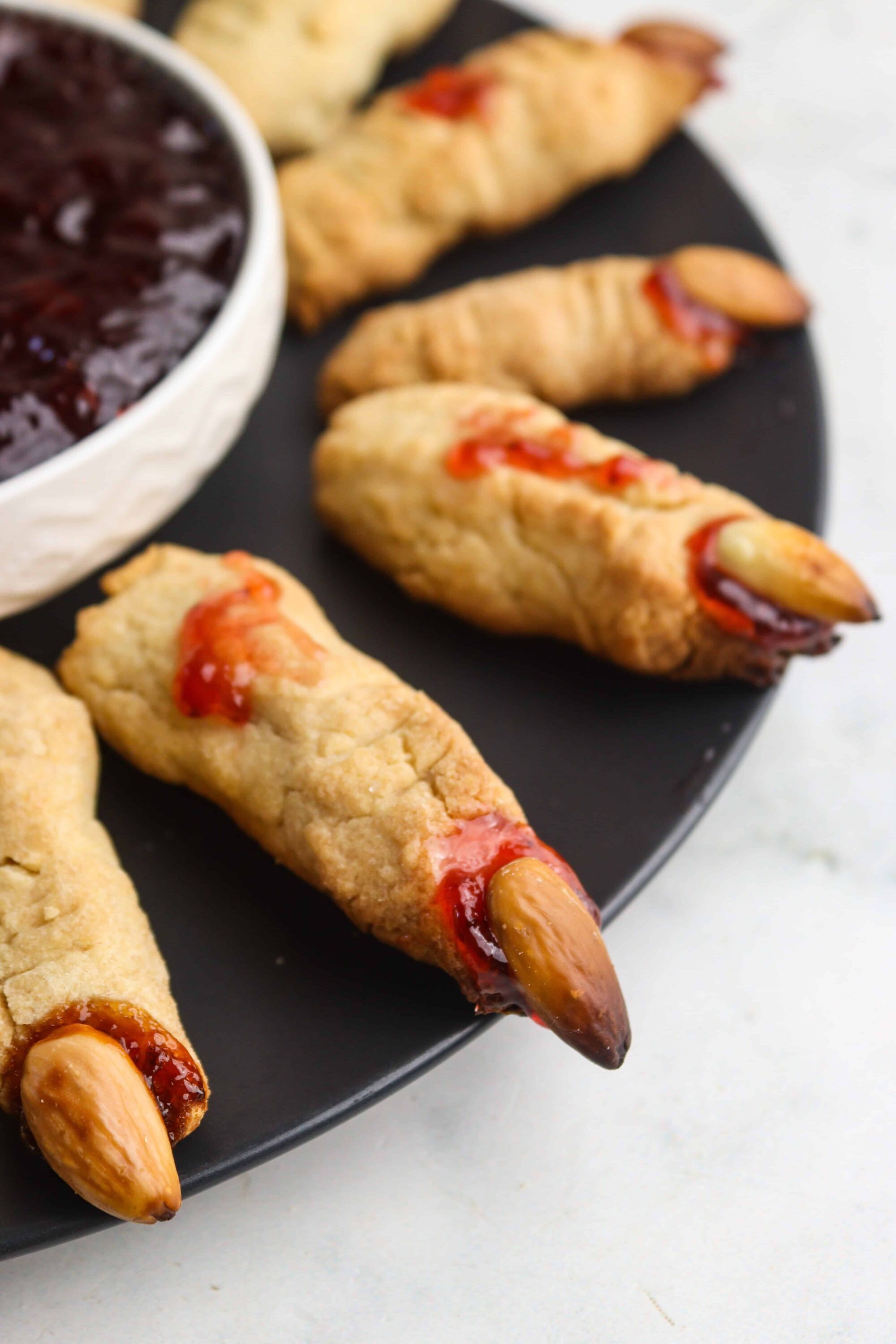Mini Halloween mummies with almond eyes on black platter, served with berry jam. Delicious, spooky treats for Halloween parties.