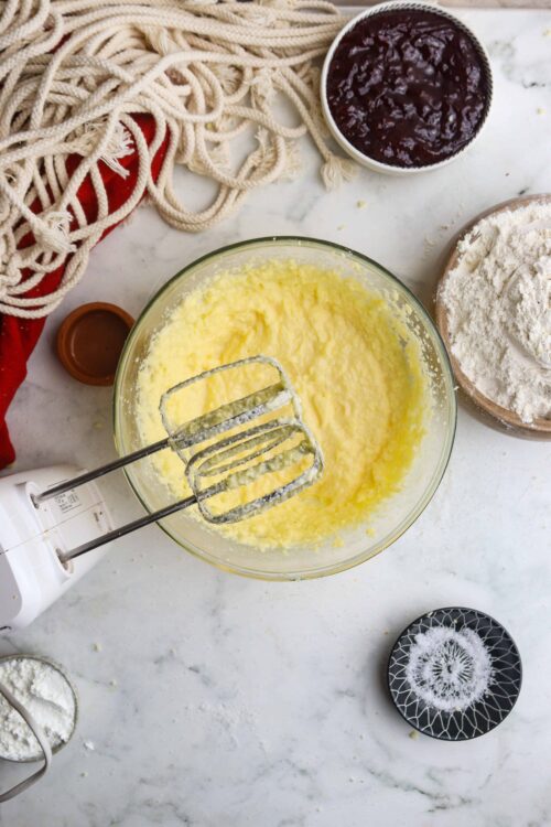 Creamy, fluffy butter mixture in mixing bowl with electric beaters, surrounded by baking ingredients and kitchen tools.