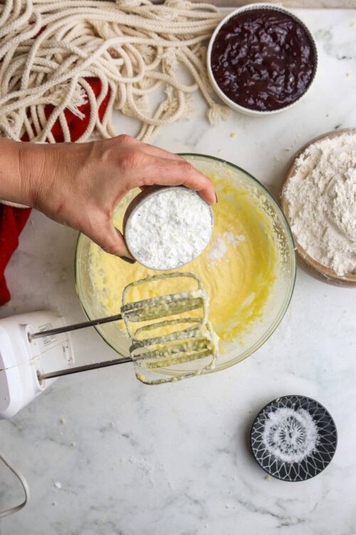 Flour being added to a mixing bowl with cake batter, surrounded by baking ingredients, on a marble countertop.