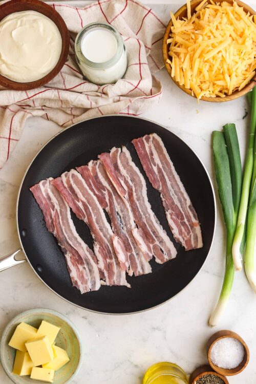 Sizzling bacon strips in a black skillet over a white marble countertop.