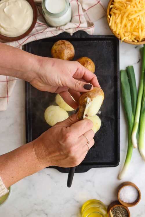 Baked potato being cut with a knife on a baking sheet with peeled potatoes in the background.