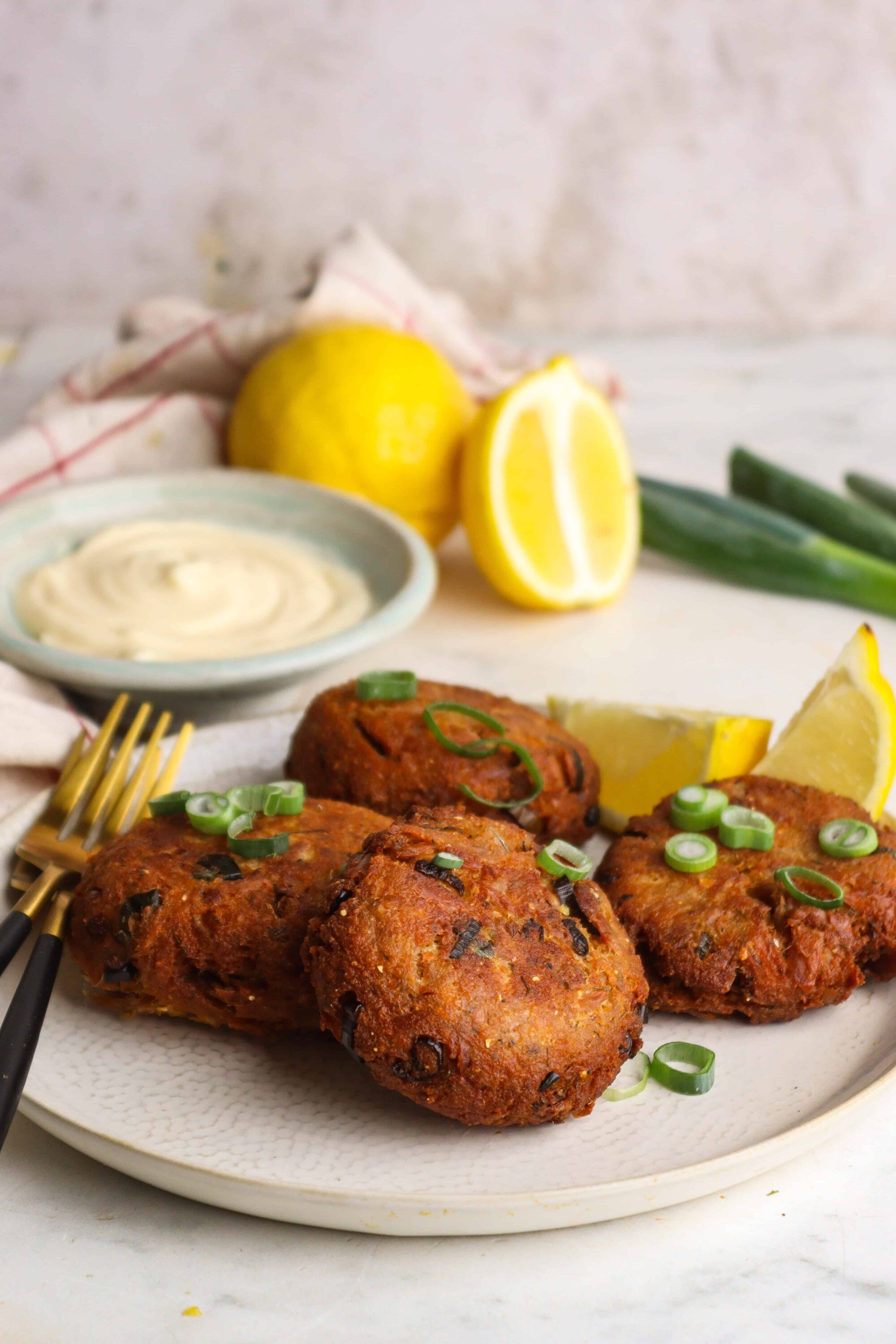 Crispy crab cakes with lemon and green onions on a white plate, served with creamy dipping sauce and fresh lemon wedges in the background.