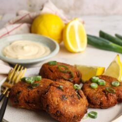 Crispy crab cakes with lemon and green onions on a white plate, served with creamy dipping sauce and fresh lemon wedges in the background.
