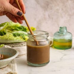 Creamy homemade salad dressing in a glass jar with a wooden spoon, fresh lettuce, and oil bottle on a marble surface.