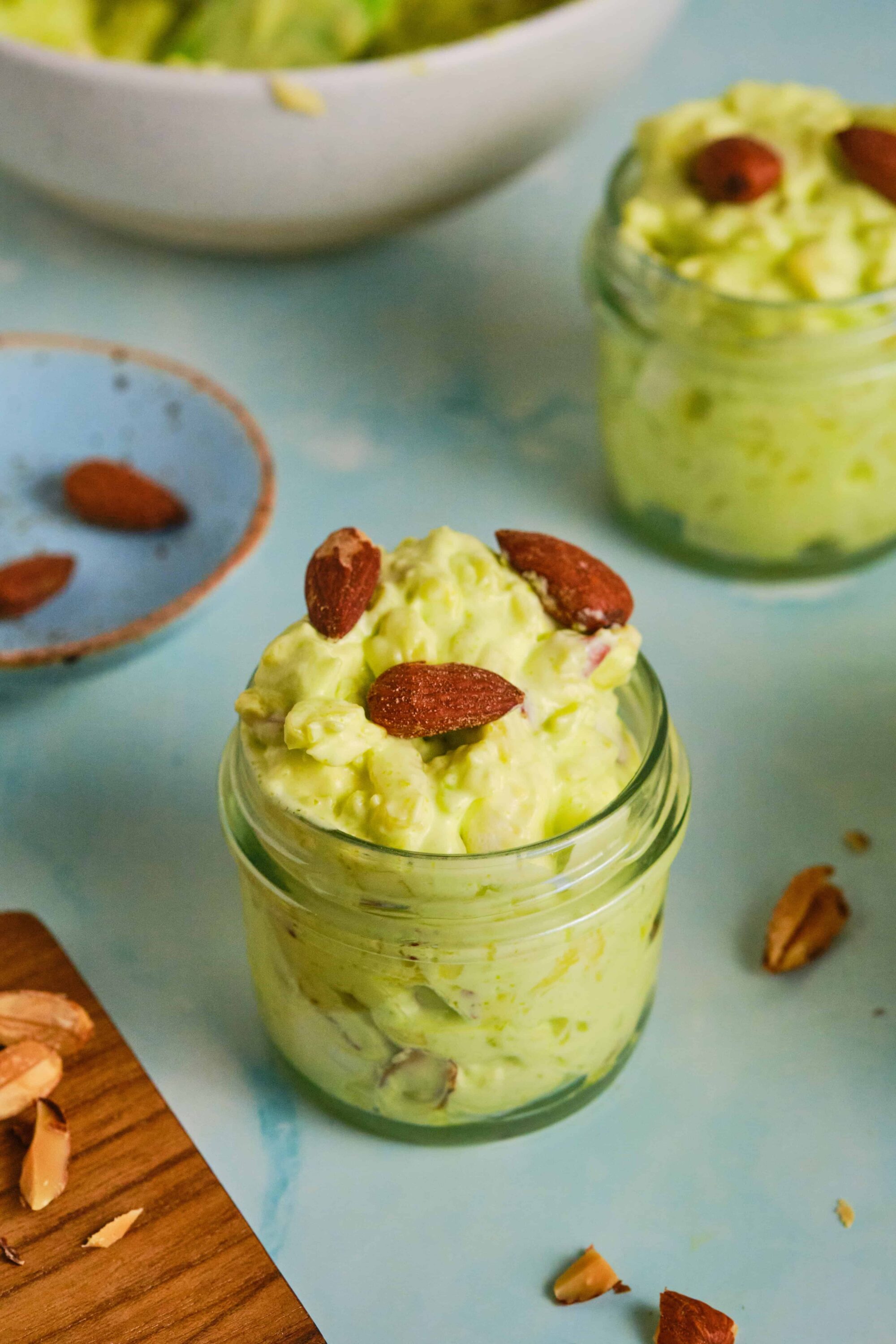 Creamy avocado and almond spread served in a glass jar, topped with whole almonds, on a light blue background.