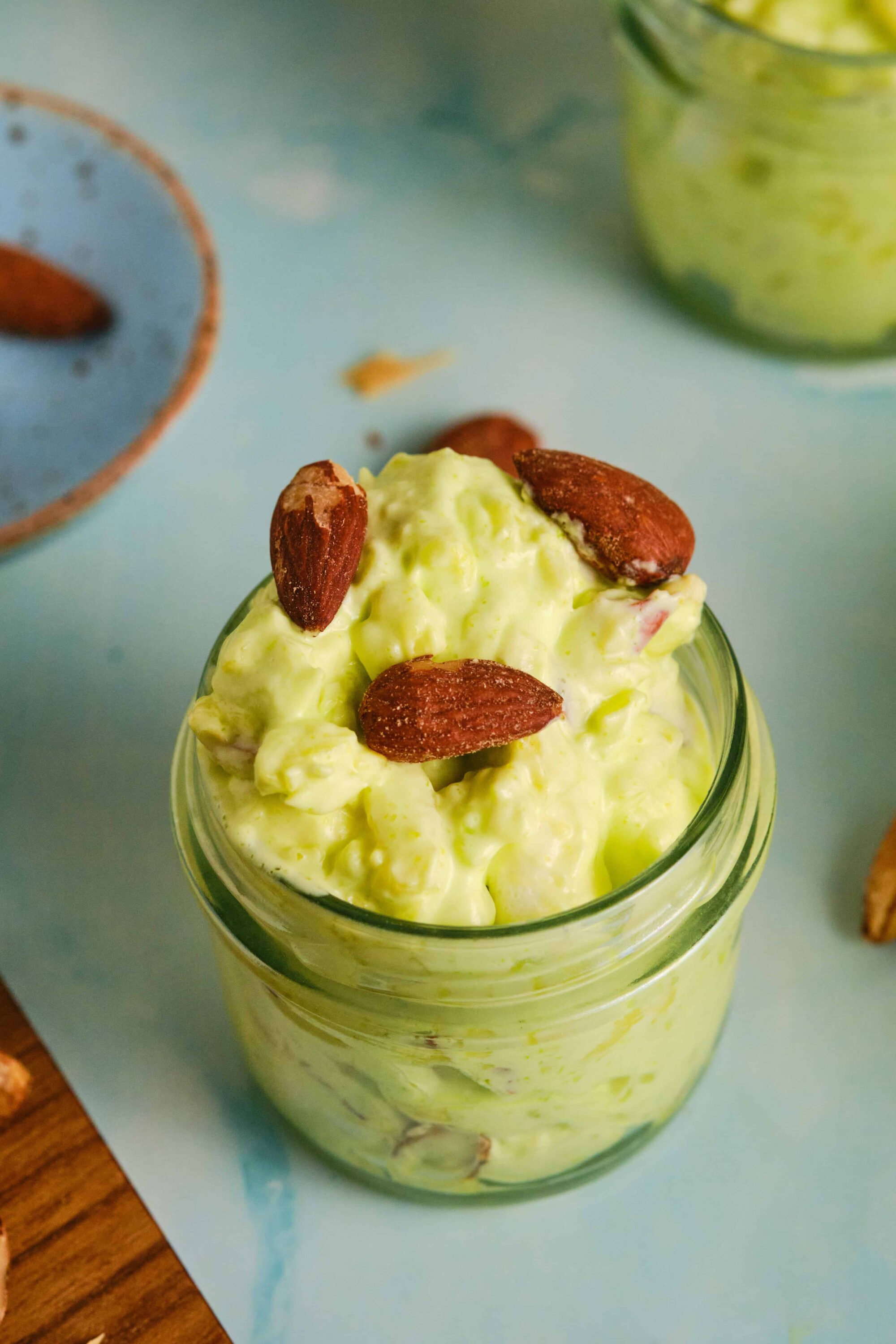 Creamy avocado and almond salad in a glass jar, topped with almonds, on a light blue surface.
